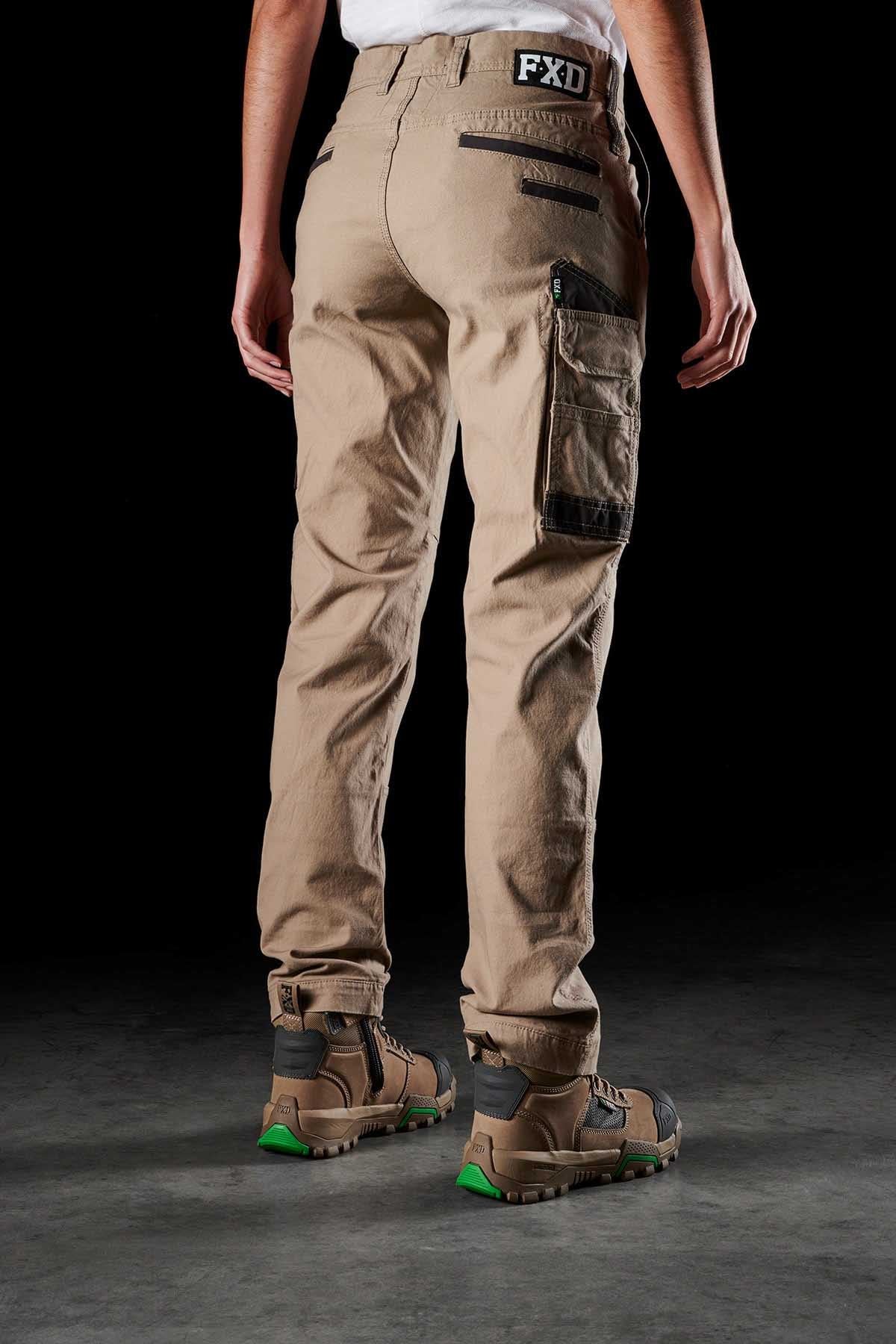 A person wearing FXD Womens WP-3W tan cargo pants with black zipper details and a brand logo on the waistband. Showcasing durable workwear, they are also wearing brown and green hiking boots. The person is standing with their back facing the camera against a dark background.