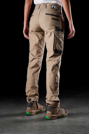 A person wearing FXD Womens WP-3W tan cargo pants with black zipper details and a brand logo on the waistband. Showcasing durable workwear, they are also wearing brown and green hiking boots. The person is standing with their back facing the camera against a dark background.