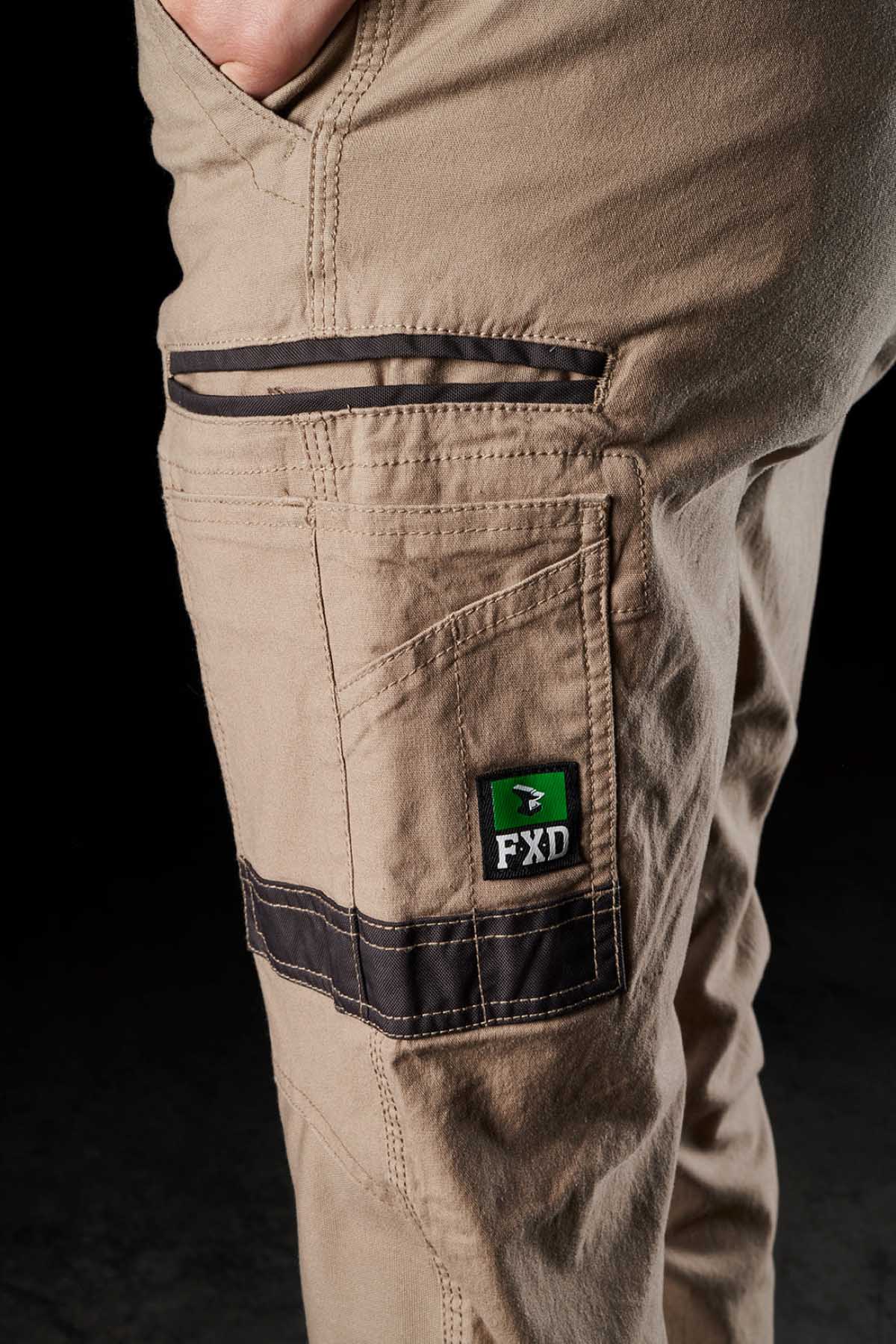 Close-up of a person wearing FXD Womens WP-3W beige work pants. The right leg features multiple pockets with black detailing, including a smaller pocket adorned with the FXD logoโa green and black patch with white lettering. The personโs hand is partially visible in the pocket, highlighting the durable workwear's 360-degree stretch designed for comfort and mobility.