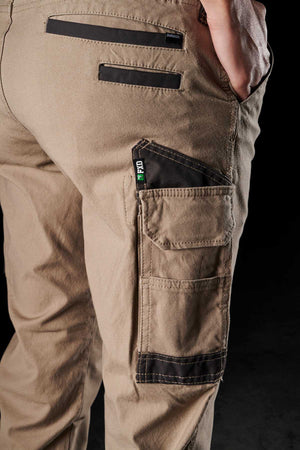 Close-up of a person wearing FXD Womens WP-3W work pants in beige, showcasing pockets and reinforced stitching. Notable details include a back pocket, a side cargo pocket with a green and black label, and durable workwear fabric with 360-degree stretch. The person's hand rests in the back pocket against a dark background.
