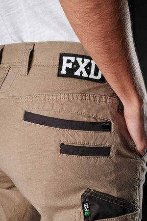 Close-up of a person wearing FXD Womens WP-3W work pants with the brand logo "FXD" on a black patch. The durable workwear features multiple pockets, including zippered ones and a small pocket with labels emphasizing durability. Thanks to the 360-degree stretch, their hand rests easily in their back pocket.