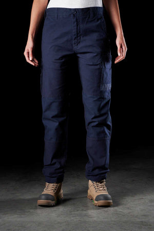 Person standing against a dark background wearing the FXD Womens WP-3W blue cargo pants with reinforced pockets and beige work boots. The photo is cropped at the neck, focusing on the lower half of the body. The women's work pant features multiple pockets on the thighs and knees.