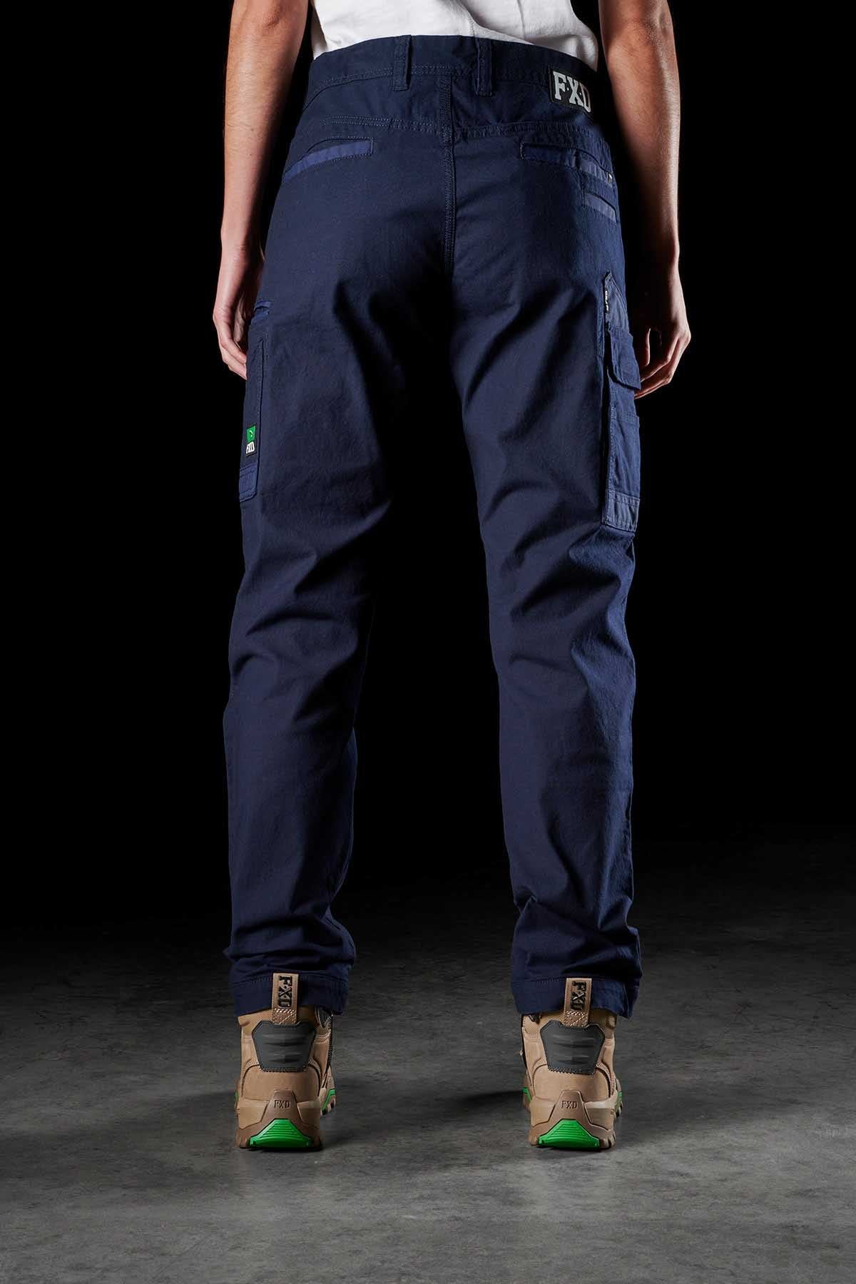 Rear view of a person wearing FXD Womens WP-3W dark blue cargo pants with 360-degree stretch and beige high-top sneakers, standing on a dark surface against a black background. The work pant features multiple reinforced pockets and a black and white patch on the waistband.