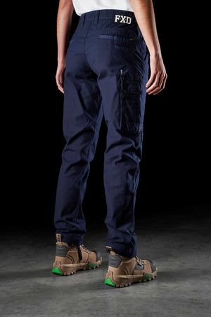A person standing with their back turned is wearing navy blue FXD Womens WP-3W work pants, which feature reinforced pockets and a "FXD" logo on the back. They are also donning beige work boots with green soles. The background consists of a black setting with a concrete floor.
