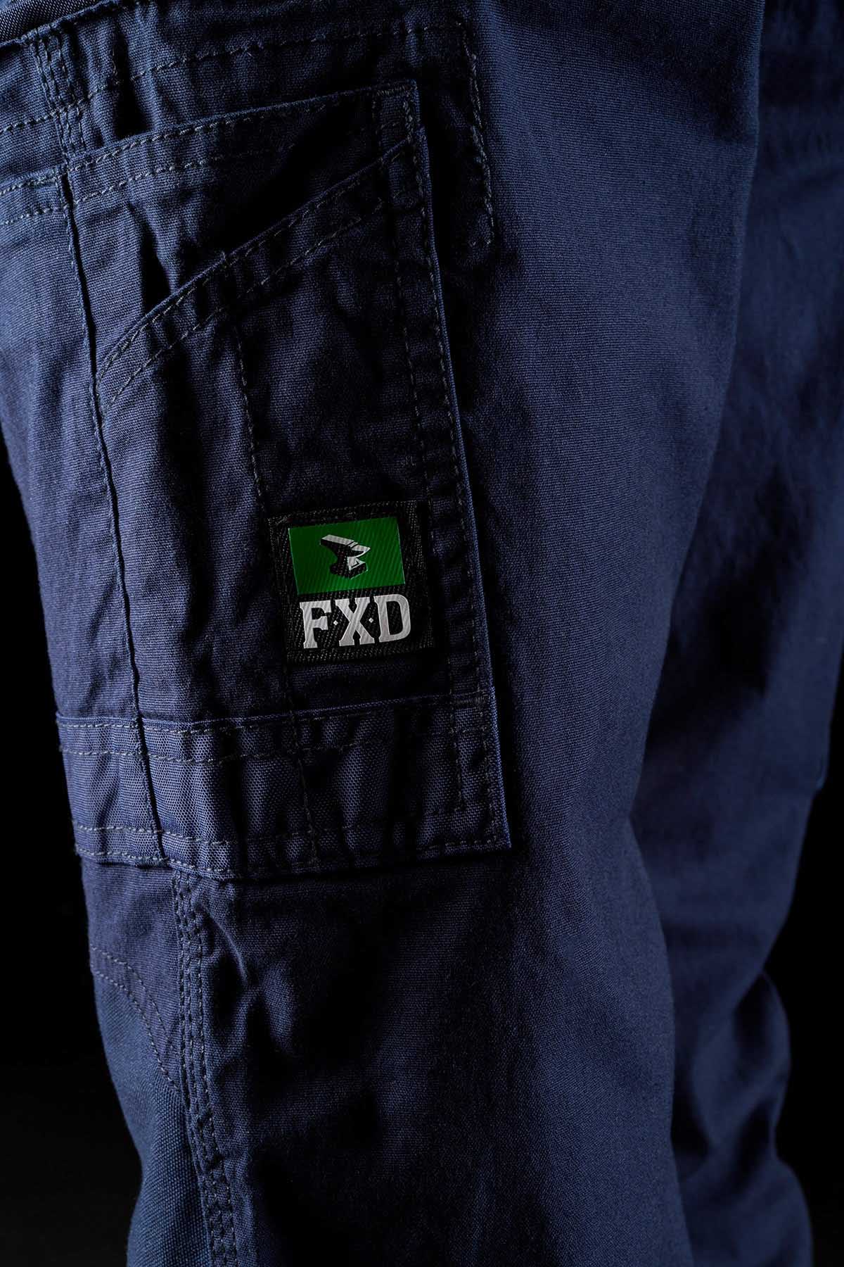 Close-up of a person wearing FXD Womens WP-3W dark blue work pants with a side pocket. The pocket features a green and black patch with the letters "FXD" and a logo of a black and white bird on a green background. The fabric appears durable and sturdy, boasting reinforced pockets for added reliability.