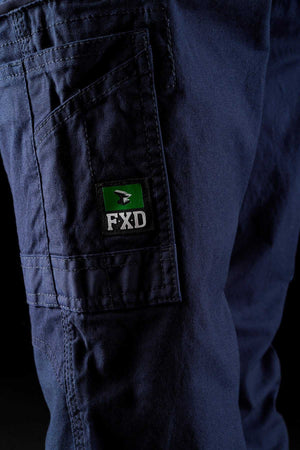 Close-up of a person wearing FXD Womens WP-3W dark blue work pants with a side pocket. The pocket features a green and black patch with the letters "FXD" and a logo of a black and white bird on a green background. The fabric appears durable and sturdy, boasting reinforced pockets for added reliability.