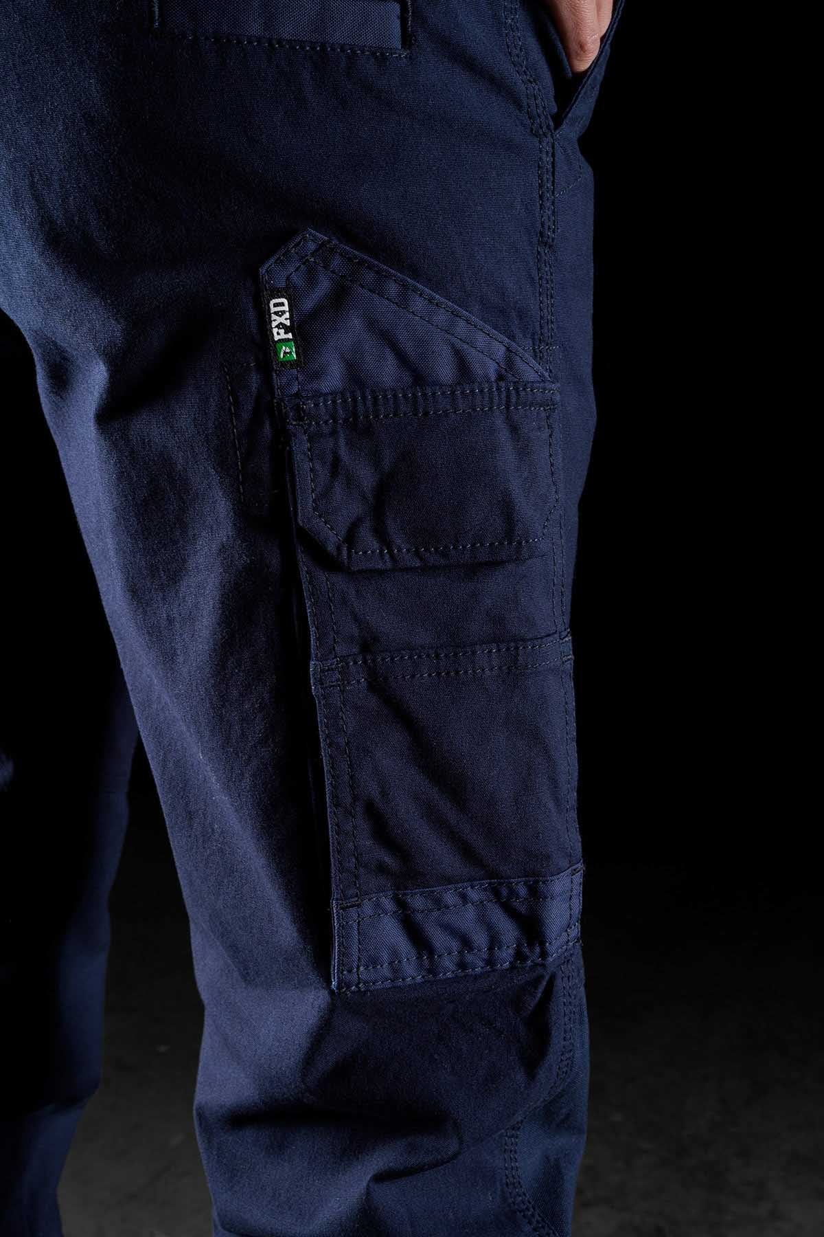 Close-up image of a person wearing FXD Womens WP-3W dark blue cargo pants with 360-degree stretch. The focus is on the left leg, showcasing multiple reinforced pockets, including a prominent angled pocket with a small green and black logo tag. The black background makes these work pants for women stand out.