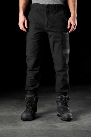 A person stands wearing FXD WP-4 Stretch Cuffed Work Pants and black work boots with green accents on the soles. Made from DURA500 Advanced Nylon and stretch cotton canvas, these pants offer durability and comfort in a sleek design. The person’s hands rest at their sides against a dark background, revealing only the lower half of their body.