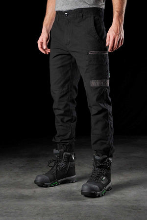 A person wearing black FXD WP-4 Stretch Cuffed Work Pants and black work boots with green accents stands against a dark background, only revealing their lower body.