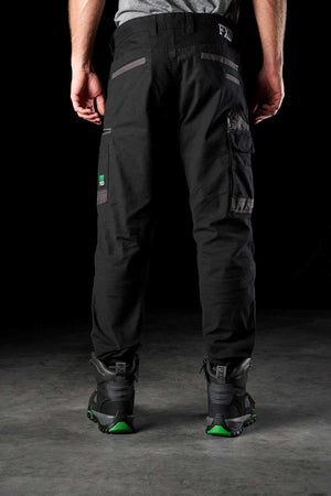 With their back facing the camera, a person is wearing black FXD WP-4 Stretch Cuffed Work Pants featuring pockets, paired with gray and green sneakers. The dark background accentuates the lower half of their body, highlighting the durability of DURA500 Advanced Nylon and stretch cotton canvas material.
