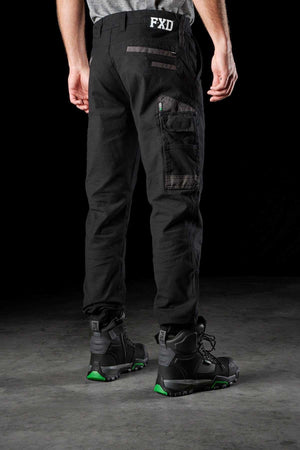 A person is standing, facing away from the camera, wearing black FXD WP-4 Stretch Cuffed Work Pants made of durable stretch cotton canvas and black work boots with green accents. The pants feature multiple pockets, and the brand "FXD" is visible on the waistband. The setting appears to be a dark, neutral space.