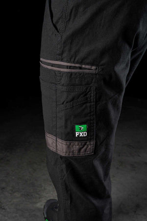 Close-up of a person's leg wearing black FXD WP-4 Stretch Cuffed Work Pants with green and grey details. Made from stretch cotton canvas, these pants feature multiple pockets, including one with a prominently displayed FXD logo. The image focuses on the side of the leg, highlighting the durable fabric and stitching.