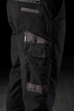 Close-up of a person wearing FXD WP-4 Stretch Cuffed Work Pants made from stretch cotton canvas with multiple pockets. The pants feature a combination of black fabric and dark grayish-brown DURA500 Advanced Nylon reinforcements on the pockets and knees. One pocket has a small label with green, white, and black colors. The background is dark.