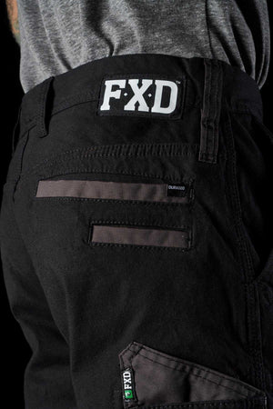 Close-up of a person wearing black FXD WP-4 Stretch Cuffed Work Pants. The pants feature a white FXD logo patch on the waistband, DURA500 Advanced Nylon black and gray pockets with the DuraEco label, and reinforced stitching. Made from stretch cotton canvas, they pair well with the gray T-shirt against the black background.