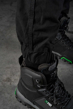 A close-up image of a person wearing black hiking boots and FXD WP-4 Stretch Cuffed Work Pants. The boots feature green accents and rugged soles, ideal for outdoor activities. The pants, crafted from stretch cotton canvas with DURA500 Advanced Nylon, are tucked in, suggesting readiness for hiking or trekking. The background is a gray, textured surface.