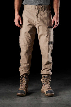 A person wearing khaki FXD WP-4 Stretch Cuffed Work Pants and brown lace-up work boots stands in front of a black background. The cargo pants feature multiple pockets and a relaxed fit. Their upper body is partially visible, showing a gray shirt.