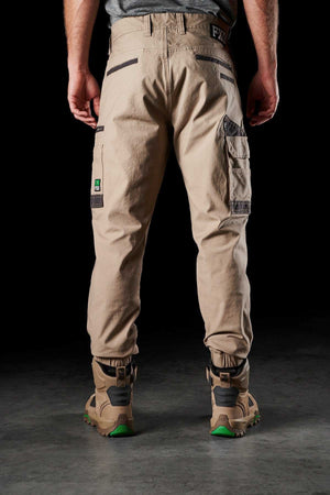 A person is standing with their back to the camera, wearing FXD WP-4 Stretch Cuffed Work Pants in beige and tan work boots with green soles. The pants, made from stretch cotton canvas, feature multiple pockets and loops against a dark background.