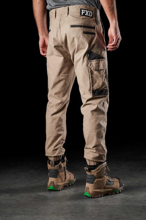A person stands facing away, wearing FXD WP-4 Stretch Cuffed Work Pants in light brown stretch cotton canvas with multiple pockets and black accents. The pants have an "FXD" logo on the back waistband. The person is also wearing rugged tan and black work boots with green soles. The background is dark.