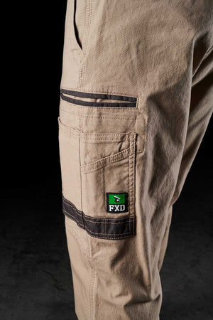 Close-up of the side pocket of light brown FXD WP-4 Stretch Cuffed Work Pants against a dark background. The pocket, crafted from stretch cotton canvas, features a black trim and an "FXD" logo with green highlights. The pants appear rugged and well-constructed, suitable for heavy-duty use.