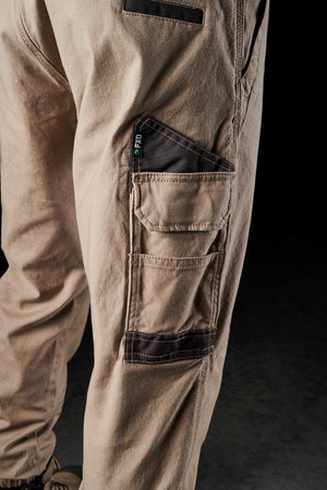 A close-up of a person wearing FXD WP-4 Stretch Cuffed Work Pants, crafted from stretch cotton canvas and featuring multiple pockets. The image emphasizes the side pocket area, showcasing details made from DURA500™ Advanced Nylon And Polyurethane Canvas, along with a visible brand tag. Only the lower part of the body is captured against a dark background.