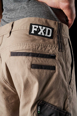Close-up of a person wearing FXD WP-4 Stretch Cuffed Work Pants in khaki, showcasing a black "FXD" logo patch on the right side. These pants are crafted from DURA500™ Advanced Nylon and Polyurethane Canvas and feature multiple pockets with dark trim and subtle stitching details. A grey shirt is partially visible at the waist.