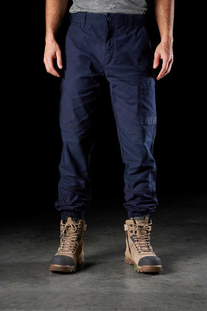 A person is standing against a black background, wearing FXD WP-4 Stretch Cuffed Work Pants made from stretch cotton canvas and tan hiking boots with green soles. The individual is dressed in a gray short-sleeved shirt, their hands by their sides. The pants feature multiple DURA500™ pockets and reinforced knees.
