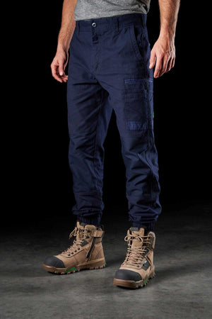 Man in a gray t-shirt showcases FXD WP-4 Stretch Cuffed Work Pants made from stretch cotton canvas, featuring multiple pockets and drawstring hems, paired with tan work boots. The outfit, enhanced with DURA500™ reinforcement, is displayed against a plain black background.