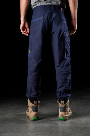 A person wearing dark blue FXD WP-4 Stretch Cuffed Work Pants and tan work boots stands with their back to the camera against a black background. The stretch cotton canvas pants, reinforced with DURA500™, have multiple pockets, including a large one on the right thigh, and the boots feature green accents on the soles.