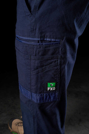 Close-up view of a person wearing FXD WP-4 Stretch Cuffed Work Pants made from stretch cotton canvas, featuring multiple pockets and an FXD logo patch. The bottom part of a brown shoe is visible against a dark background.