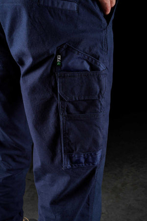 Close-up image of a person wearing dark blue FXD WP-4 Stretch Cuffed Work Pants made from stretch cotton canvas with multiple pockets. The person has one hand partially visible and appears to be standing against a dark background. The pockets have flaps, detailed stitching, and feature DURA500™ reinforcement.