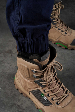 A close-up view of a person wearing tan hiking boots with thick soles and green accents. The individual is dressed in dark blue FXD WP-4 Stretch Cuffed Work Pants, with the trousers tucked into the boots made from stretch cotton canvas. The image is taken from ground level on a gray concrete surface.