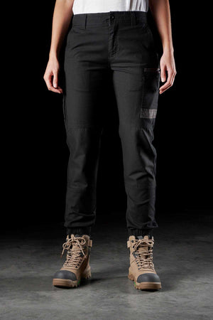 A person wearing FXD Womens WP-4W black stretch work pants with DURA500 reinforced pockets and brown hiking boots stands against a dark background. Their hands are relaxed by their sides, showcasing the multiple pockets on the Cotton Twill cargo pants, while the hiking boots are laced up securely.