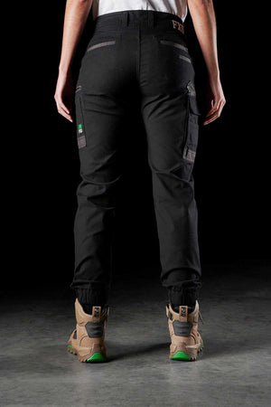 A woman wearing FXD Womens WP-4W black stretch work pants with DURA500 reinforced pockets and beige boots featuring green accents stands facing away against a dark background, with only her lower back and legs visible.