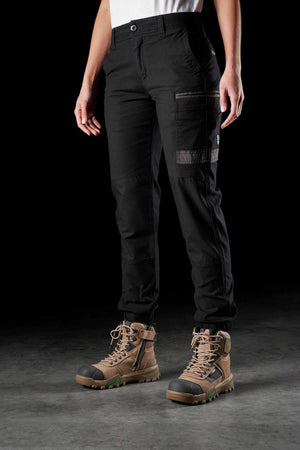 A person stands against a dark background, wearing FXD Womens WP-4W black cargo pants with DURA500 reinforced pockets and tan hiking boots with black toe caps and green accents. The pants, made from Cotton Twill and featuring flap pockets on the thighs, allow for durability and flexibility as the person's hands rest by their sides.