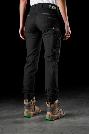 A person wearing FXD Womens WP-4W black stretch work pants with DURA500 reinforced back and side pockets, paired with brown ankle-high work boots featuring green soles, stands on a dark surface. The "FXD" logo is visible on the waistband at the back of the pants as the person faces away from the camera.