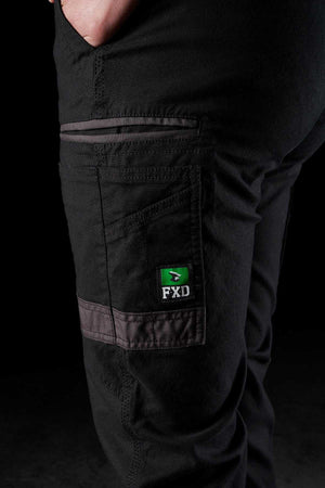 A close-up shows someone wearing FXD Womens WP-4W black stretch work pants with a hand in the side pocket. The "FXD" logo is visible on a pocket flap. These cotton twill trousers feature reinforced stitching, grey trim details, and DURA500 reinforced pockets against a black background.