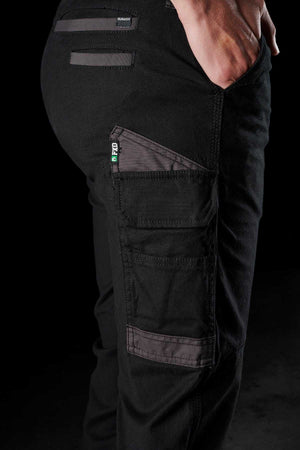 Close-up of a person wearing FXD Womens WP-4W black stretch work pants with their hand tucked into one of the DURA500 reinforced pockets. The pants feature several utility pockets, including a prominent side pocket with a small green, white, and black logo tag. The background is dark, highlighting the pants.