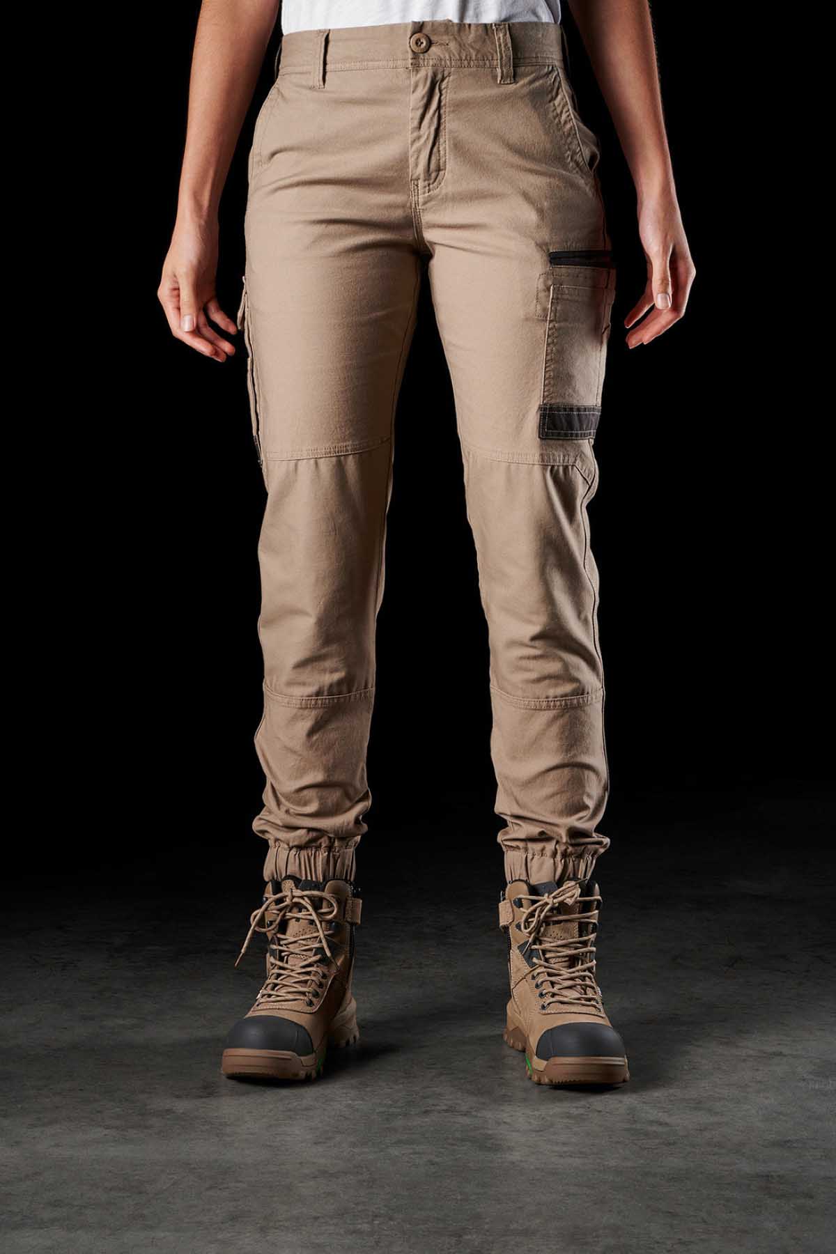 A person standing against a dark background is wearing FXD Womens WP-4W beige cotton twill cargo pants with multiple DURA500 reinforced pockets and brown lace-up boots. The pants are gathered at the ankles, and the person's hands are hanging by their sides.