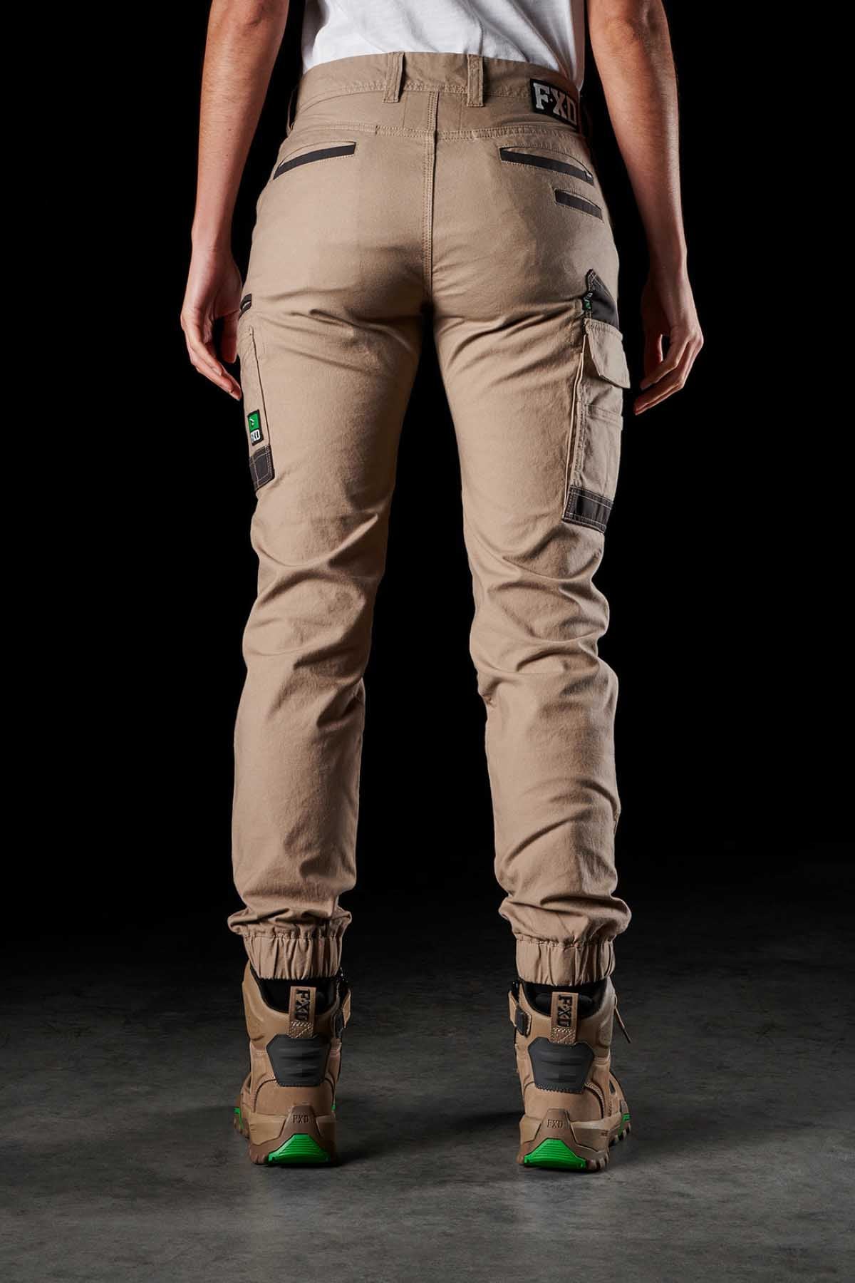 A person is standing against a dark background, facing away from the camera, wearing FXD Womens WP-4W beige stretch work pants with DURA500 reinforced pockets and tan work boots featuring green accents. The pants have multiple pockets and details, while the person sports a tucked-in white shirt.