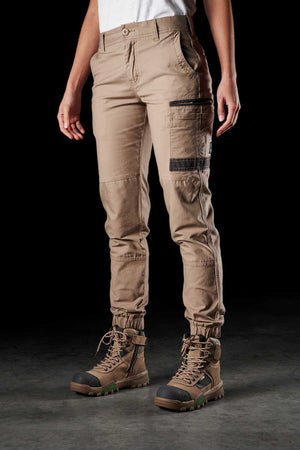 A person wearing the FXD Womens WP-4W beige utility pants with multiple pockets and green-soled hiking boots stands against a dark background. The Cotton Twill pants feature reinforced knee areas and ankle cuffs for durability. Their upper body remains out of view.