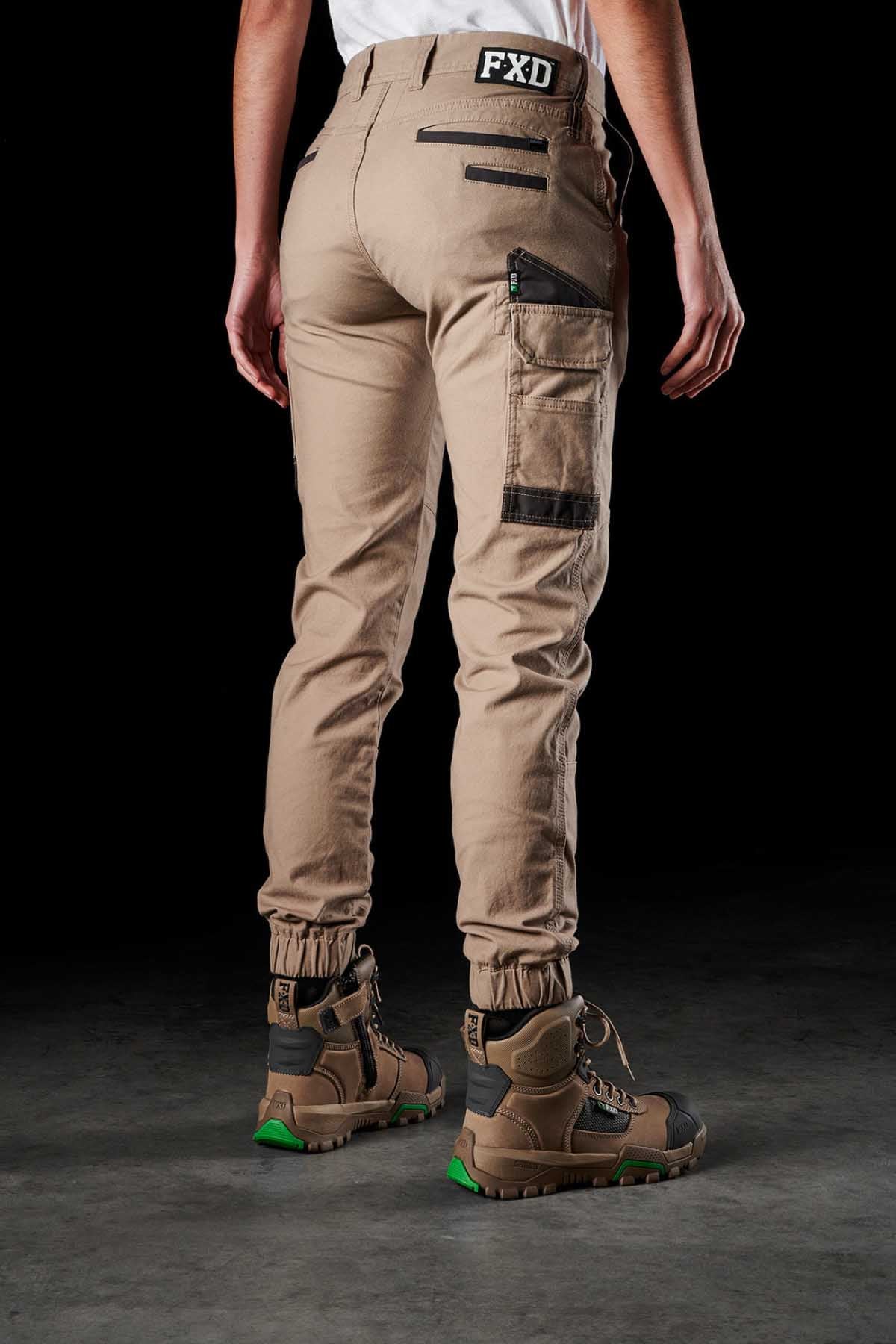 A person is standing with their back facing the camera, wearing FXD Womens WP-4W tan cargo pants made from durable cotton twill with black zipper details and a white top. The pants are cinched at the ankles and complemented by brown and green hiking boots. The background is black.