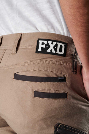 Close-up of the back of a person wearing FXD Womens WP-4W tan stretch work pants with a white T-shirt tucked in. The cotton twill pants feature black trim on the back pockets, DURA500 reinforced pockets, and a black label on the waistband with "FXD" written in white letters. The person has their right hand in their pocket.