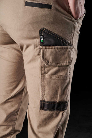 Close-up view of a person wearing FXD Womens WP-4W tan cargo pants with black accents. These stretch work pants feature DURA500 reinforced pockets, including a buttoned pocket and a zippered one, with a visible green and white "WTFD" tag. The background is black.