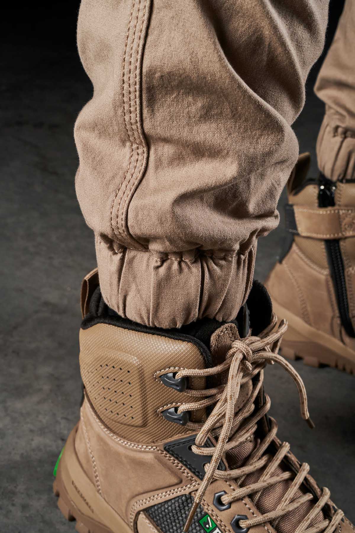 A close-up of a person wearing FXD Womens WP-4W tan, lace-up work boots with green accents. The stretch work pant is tucked into the boots and secured with an elastic band around the ankle. DURA500 reinforced pockets add to the durability, while the background reveals a concrete floor.