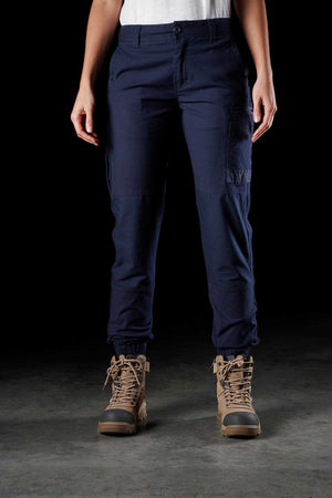 A person stands against a dark background, wearing the FXD Womens WP-4W navy blue cargo pants made from durable Cotton Twill with reinforced pockets and brown hiking boots. The person's torso and head are not visible, while their hands rest by their sides. The lighting highlights the details of the pants and boots.