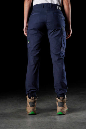 A person wearing the FXD Women's WP-4W navy blue cargo pants and tan work boots stands on a gray surface against a black background. The stretch work pants, made of durable Cotton Twill, feature multiple reinforced pockets with two green labels visible—one on the back pocket and another on the side pocket.