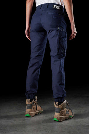 A person in a white T-shirt is shown from the back, wearing navy blue FXD Womens WP-4W cargo pants made of durable Cotton Twill with multiple reinforced pockets and a black "FXD" label on the waistband. They are also wearing brown work boots with green accents. The background is black.