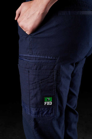 A person wearing dark blue FXD Womens WP-4W stretch work pants is shown from the waist down, with their left hand placed in the back pocket. The pants feature reinforced pockets and a green and black FXD logo on the side pocket. The background is black.
