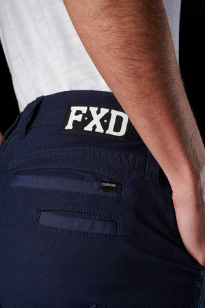 Close-up of a person wearing FXD Womens WP-4W navy blue stretch work pants with a patch that reads "FXD" on the back waistband. The person's hand is partially visible on the right side. The cotton twill pants have reinforced rear pockets, one featuring a "DURAS500" label. The background is black.