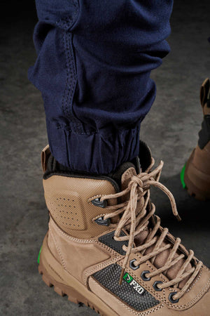 A close-up of a person wearing FXD Womens WP-4W blue cargo pants, crafted from durable Cotton Twill, tucked into tan protective work boots with green accents. The boots feature sturdy laces and a rugged design suitable for industrial or outdoor environments. The person stands on a gray, textured surface.